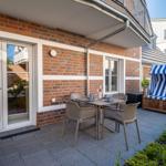 Terrasse mit Tisch, Stühlen und Strandkorb. Hintergrund: Backsteinmauer und Glas-Tür.