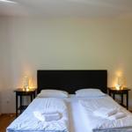 Schlafzimmer mit Doppelbett, zwei Nachttischen und Stuhl. Wandbild und Vorhänge sind sichtbar.