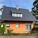 Ein orangefarbenes Haus mit dunklem Dach und Fenstern. Umgeben von Bäumen und einem gepflasterten Weg.