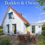 Ferienhaus mit Terrasse, Tisch und Stühlen. Roter Dachfirst, weiße Fassade und grüne Umgebung.
