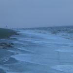 Strand mit Wellen, die an den Uferbereich schlagen, und grüner Hang im Hintergrund.