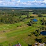 Luftbild eines Golfplatzes mit grünen Fairways, Bäumen und Teichen. Im Hintergrund sind Wälder und ein See sichtbar.