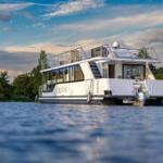 Ein weißes Hausboot mit Balkon liegt auf ruhigem Wasser zwischen Bäumen.