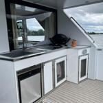 Außenküche mit Spüle, Grill und Kühlschrank an Bord. Blick auf Wasser und Ufer.