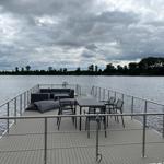 Terrasse mit Sitzgruppe und Tisch über Wasser. Himmel bewölkt, Ufer im Hintergrund.