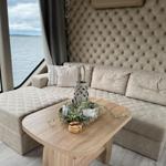 Sofa mit Hocker und Tisch vor großem Fenster mit Blick auf Wasser.