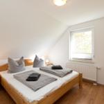 Schlafzimmer mit Holzbett, Nachttisch und Fenster. Holzfußboden und weiße Wände.