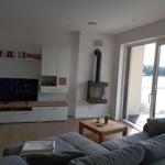 Wohnzimmer mit Sofa, Fernseher und Kaminofen. Durchgang zur Terrasse mit Blick auf Wasser.
