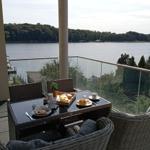 Terrasse mit Tisch und Stühlen, Blick auf einen See mit Wald im Hintergrund.