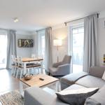 Wohn- und Essbereich mit Sofa, Esstisch und Küchenzeile. Große Fenster und Türen.