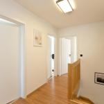 Flur mit Holzboden, weißen Türen und Holzgeländer. An der Wand hängen zwei Bilder.