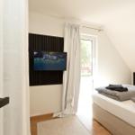 Schlafzimmer mit Bett, TV und Fenster. Holzboden und weiße Wände.