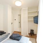 Schlafzimmer mit Bett, Kleiderschrank und Fernseher. Fenster mit weißen Vorhängen.