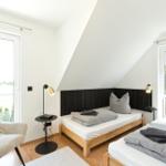 Zwei Betten mit Holzrahmen, schwarze Kopfteilwand, zwei Fenster und ein Sessel.