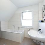 Badezimmer mit Badewanne, Waschbecken und Fenster. Weiße Fliesen und schwarze Handtuchhalter.