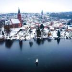 Schneebedeckter Ort mit Kirche und Häusern am Wasser, ein Segelboot fährt auf dem See.