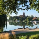 Seeufer mit Bänken, Kirche und Booten im Hintergrund.