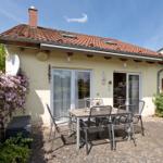Haus mit Terrasse, Garten und Tisch mit Stühlen. Dach mit Schornstein und Fenstern.