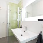 Badezimmer mit Dusche, Waschbecken und Spiegel. Wandfliesen in Grün und Weiß.