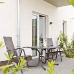 Terrasse mit Tisch und zwei Stühlen, umgeben von Pflanzen und einer Glastür.