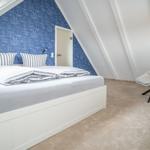 Bedroom with sloped ceilings, blue sailboat wallpaper, white bed, and chair.