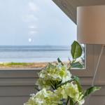 Through the window you can see the sea. In the foreground, there is a lamp and a flower vase.