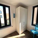 Schlafzimmer mit weißem Schrank, blauer Stuhl und zwei Fenstern mit Blick auf Gebäude.