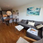 Wohnzimmer mit Sofa, Esstisch und Küchenbereich. Wandbild zeigt Meereslandschaft.
