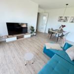 Wohn- und Essbereich mit Sofa, TV und Tisch. Holzboden und weiße Wände.