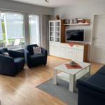 Wohnzimmer mit blauen Sesseln, weißem Couchtisch und TV-Schrank. Holzboden und große Fenster.