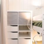 Ein weißes Bett mit grünem Bettzeug neben einem Schrank mit Fächern und einer Wandlampe.
