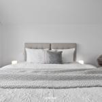 Doppelbett mit Kopfteil, Bettwäsche und Decke. Beleuchtung und Sessel sind sichtbar.