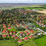 Luftbild eines Dorfes mit roten Dächern, grünen Wiesen und einem Hafen im Hintergrund.