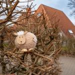 Ein hölzernes Schaf mit Wolle sitzt auf Ästen vor einem Haus mit rotem Dach.