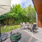 Terrasse mit Tisch, Stühlen, Grill und Sonnenschirm. Grünflächen und Bäume im Hintergrund.