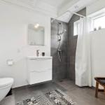 Badezimmer mit Dusche, Waschbecken und Toilette. Weiße Wände, graue Fliesen und Holzstuhl.