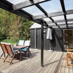 Terrasse mit Tisch, Stühlen und Sitzbank unter Dach. Hintergrund: grüner Garten und Haus.