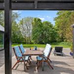 Terrasse mit Tisch, vier Stühlen und blauen Kissen. Hintergrund: grüner Garten und Bäume.