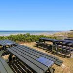 Drei Picknicktische stehen auf einer Wiese mit Blick auf den Strand und das Meer.