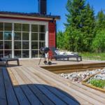 Holzterrasse mit Liegen und Grill vor einem roten Haus mit großem Fenster.