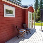 Roter Holzhaus mit Terrasse, Stühlen und Sonnenschirm. Hinterhof mit Grünfläche und Bäumen.
