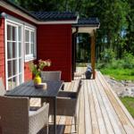Roter Holzausbau mit Terrasse, Tisch und Stühlen. Hintergrund: Wald und Grünfläche.