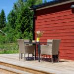 Holzterrasse mit Tisch und Stühlen vor rotem Holzhaus. Grünflächen und Bäume im Hintergrund.