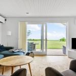 Wohnzimmer mit Sofa, Tisch und TV. Durchgehende Glastür führt auf eine Terrasse mit Blick auf Grasland.