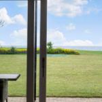 Von der Terrasse aus Blick auf grünen Rasen, gelbe Blumen und das Meer unter blauem Himmel.