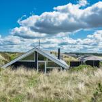 Moderne Hütte mit Dachgaube, umgeben von Gras und Bäumen unter blauem Himmel mit Wolken.