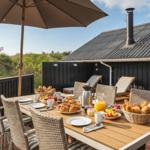 Terrasse mit Esstisch, Sonnenschirm und Liegestühlen. Frühstückstablett mit Brot, Obst und Getränken.