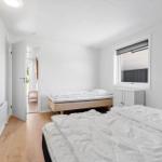 Schlafzimmer mit zwei Betten, Holzboden und Fenster. Tür führt in einen Flur.