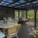 Terrasse mit Holztisch, Stühlen und Sitzbank unter Dach. Grün hinter Glas.