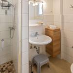 Badezimmer mit Dusche, Waschbecken und Toilette. Holzschrank und Spiegel sichtbar.